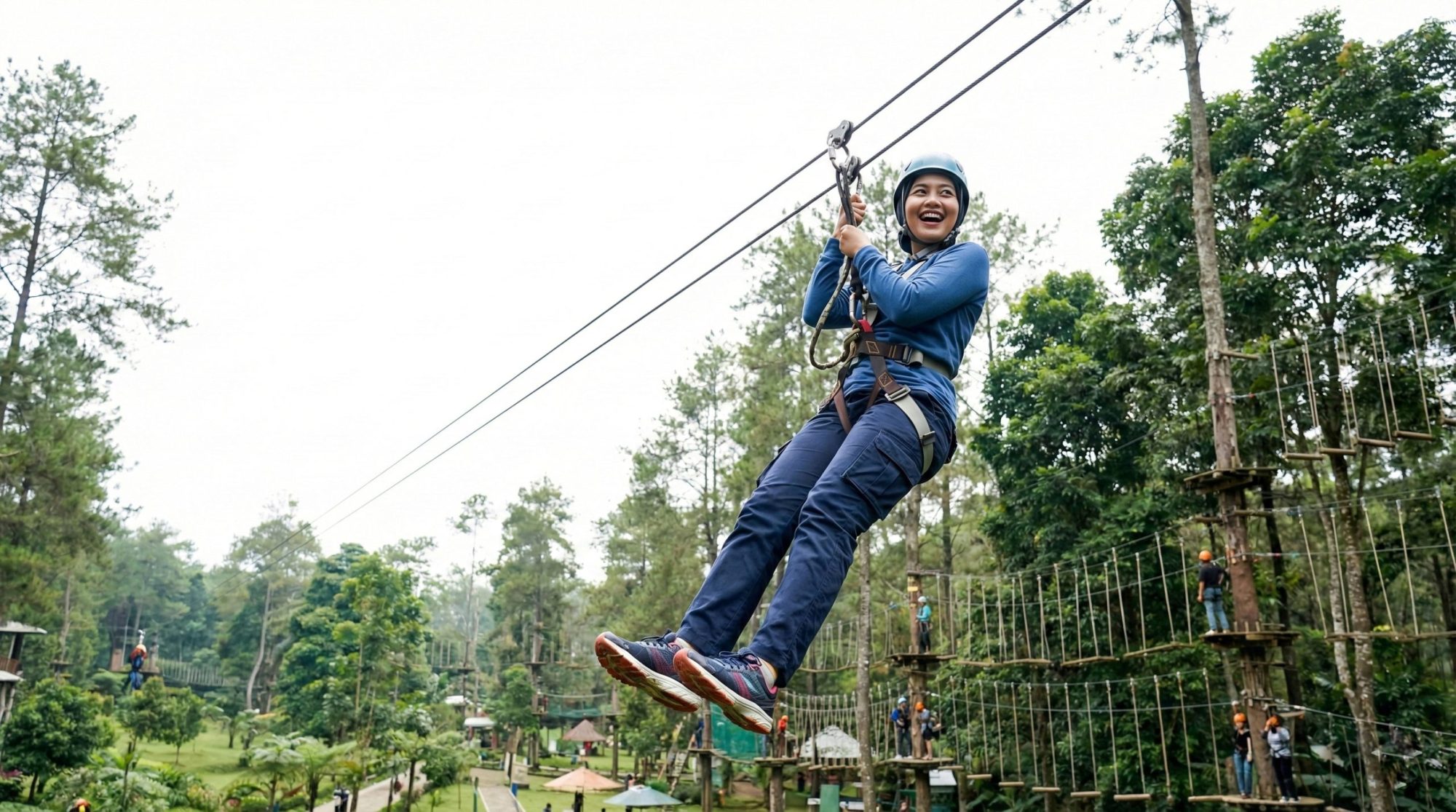 Peserta outbound Indonesia meluncur di lintasan flying fox dari ketinggian, memakai helm dan harness, dengan latar pepohonan dan suasana alam terbuka.