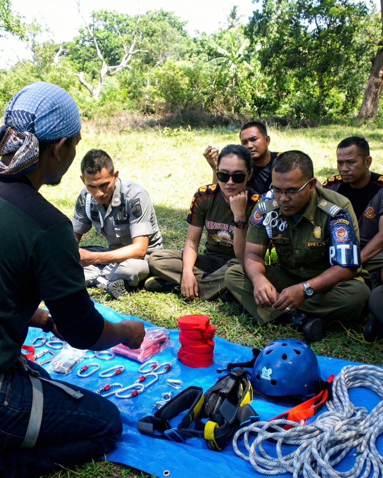 Peserta diklat lembaga sedang menerima pengarahan outdoor, duduk bersama instruktur dengan peralatan keselamatan di depannya.